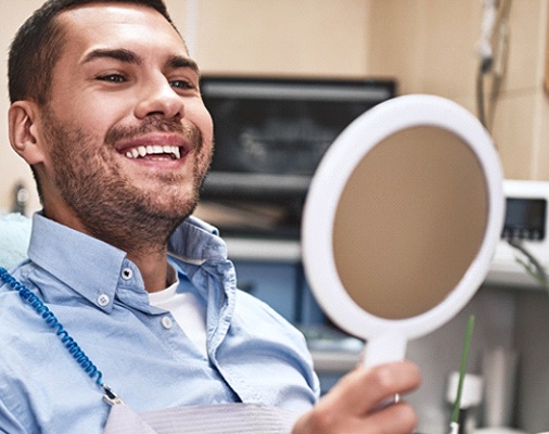 Man smiling in dental chair while looking at handheld mirror